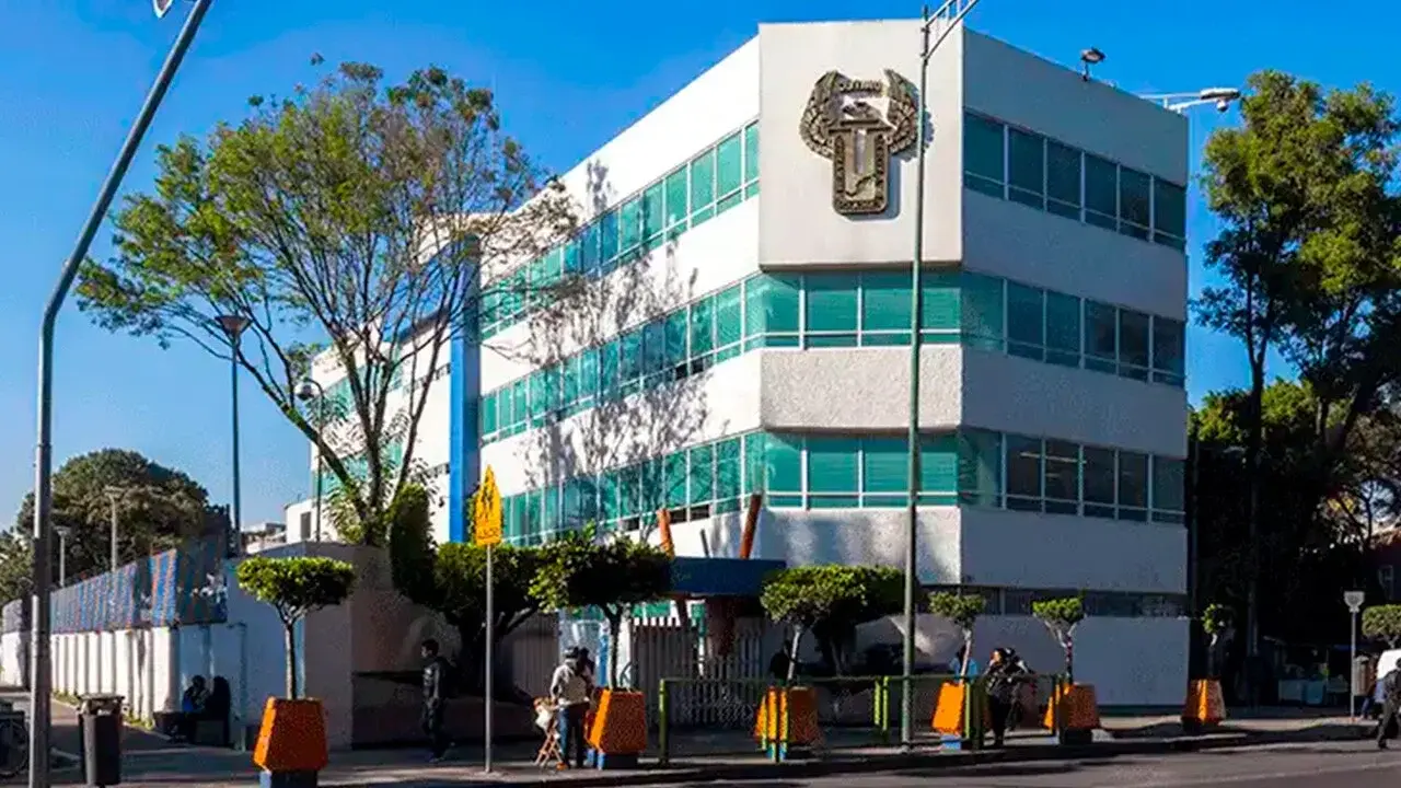 Un edificio de oficinas de varios pisos, de color blanco con detalles verdes, se encuentra en una calle urbana arbolada.