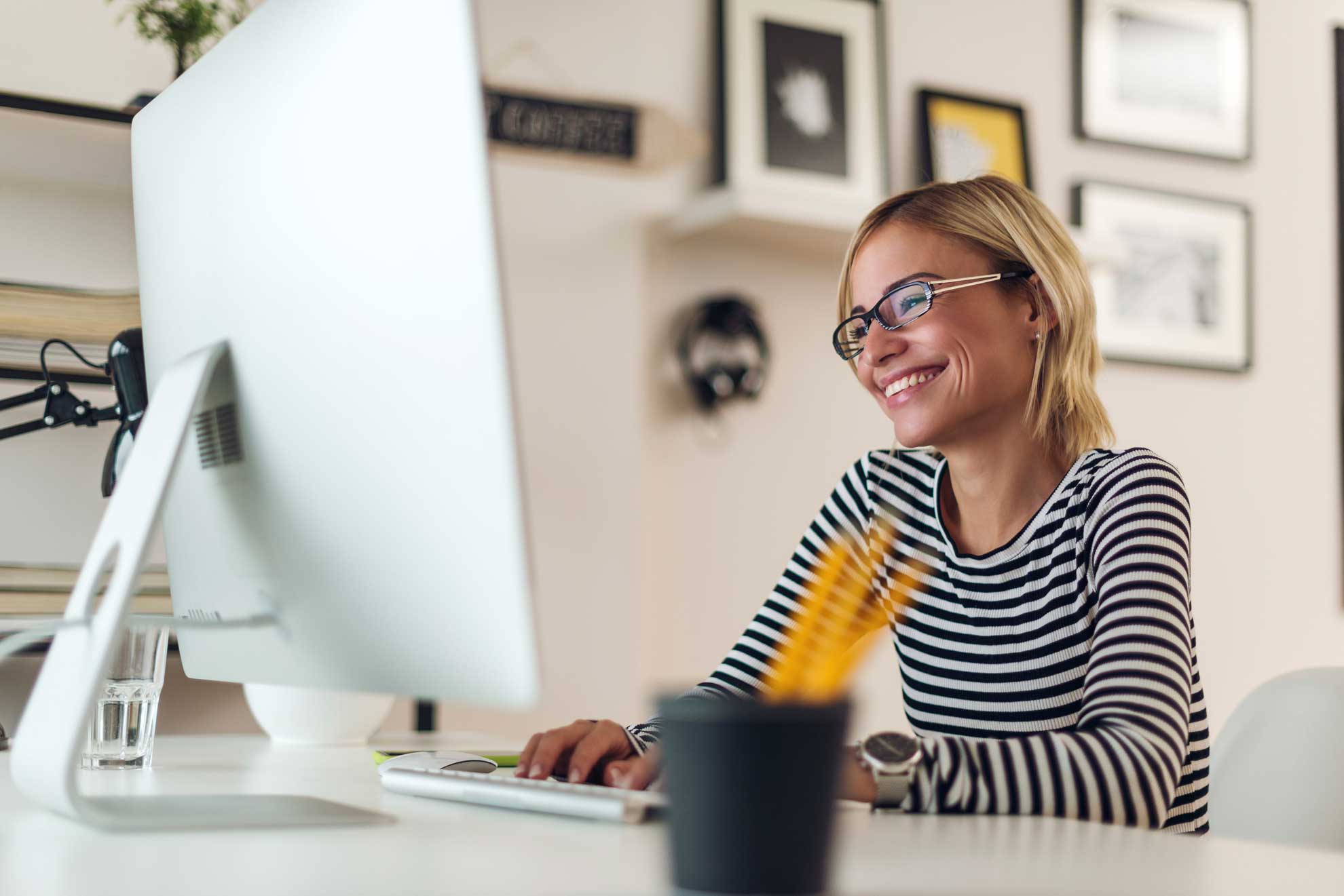 Mujer sonriente trabajando frente a un monitor de escritorio en una oficina decorada y moderna.